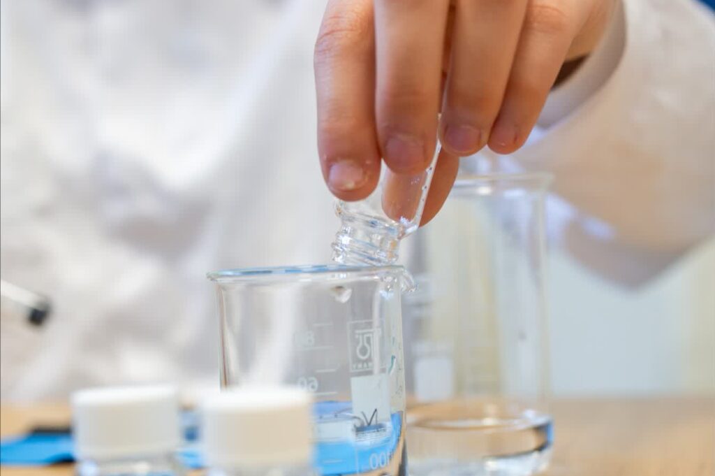 Uczennica w białym fartuchu laboratoryjnym pipetuje płyn do zlewki, na stole stoją probówki z kolorowymi cieczami.